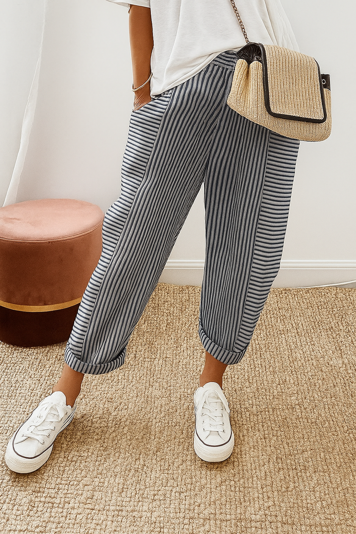Person wearing navy blue striped pants with white sneakers and a casual white top, standing on a textured beige carpet near a brown stool.