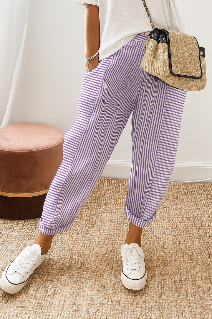 Person wearing purple striped pants with white sneakers and a casual white top, standing on a textured beige carpet near a brown stool.