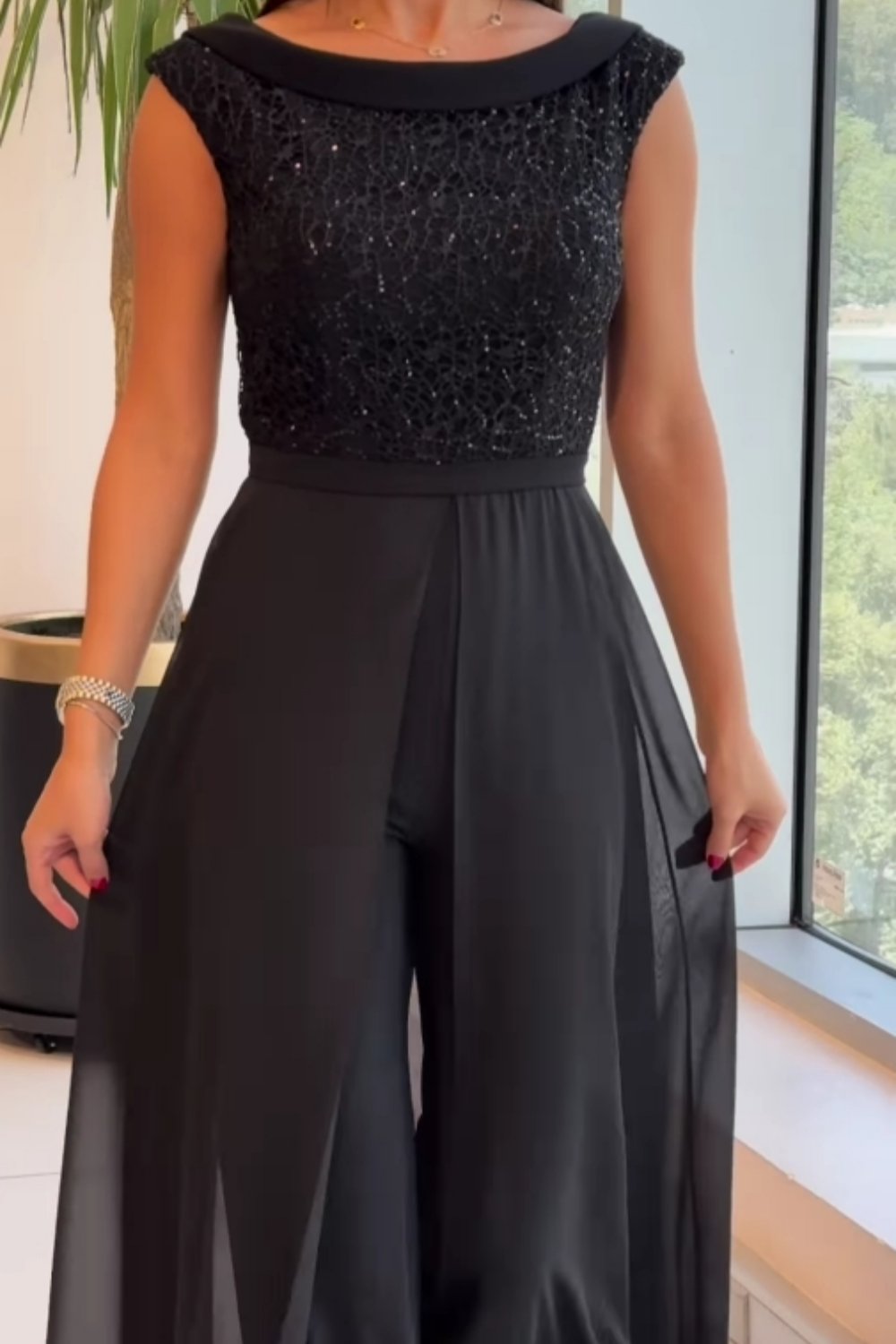 Close-up of a black textured tulle jumpsuit with sheer flowing pants, worn by woman in an indoor setting near a large window.
