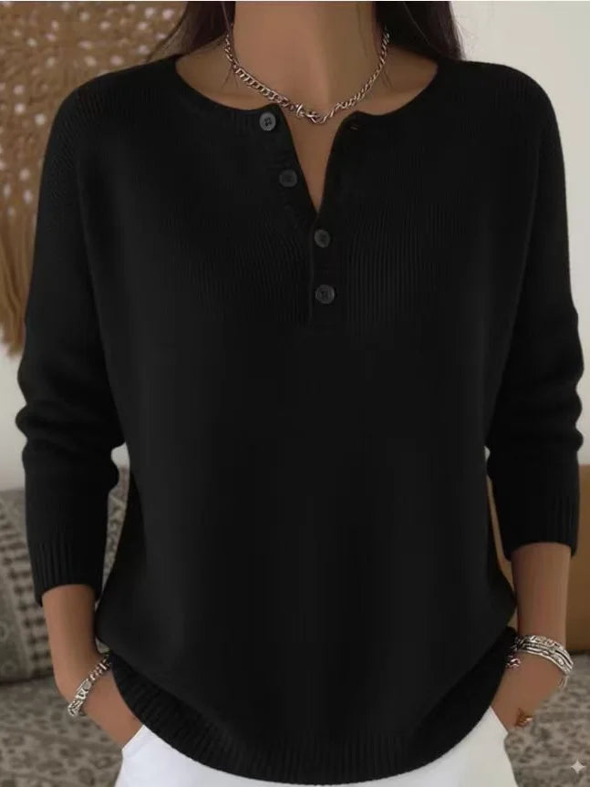 Model wearing black sweater with buttoned neckline, white jeans, bracelets, indoors with patterned cushions.