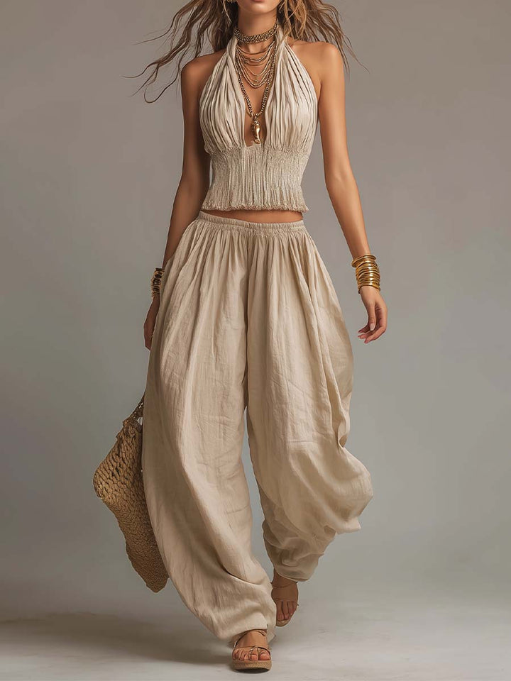Beige two-piece set with a halter top and billowy pants, worn with jewelry and sandals. Studio background.