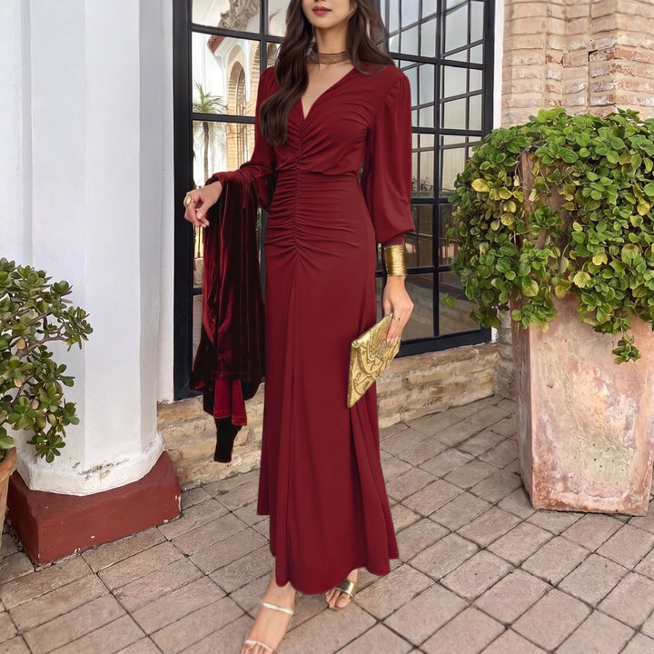 Elegant burgundy midi dress with ruched detail and long sleeves, styled with gold accessories, in an outdoor patio setting.