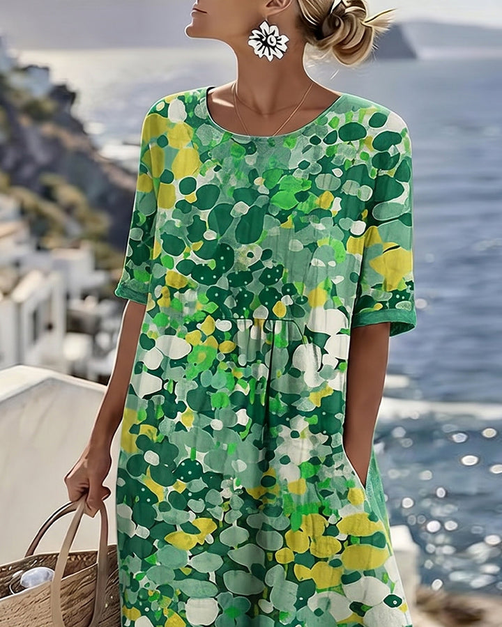 Model in blue and green floral dress on seaside terrace, holding bag with ocean view.