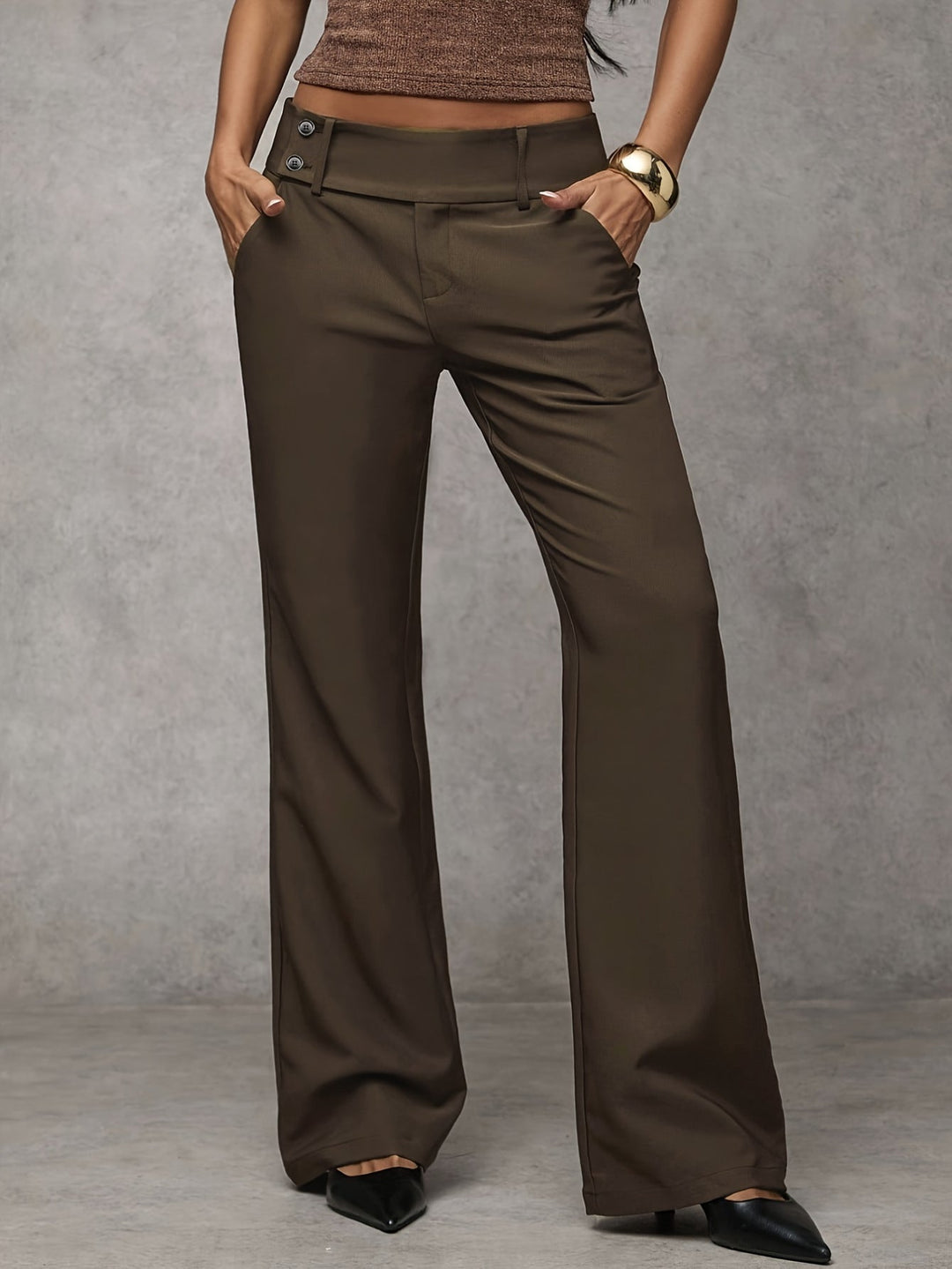 Brown modern pants with button detail, modeled with hands in pockets. Indoor gray background.