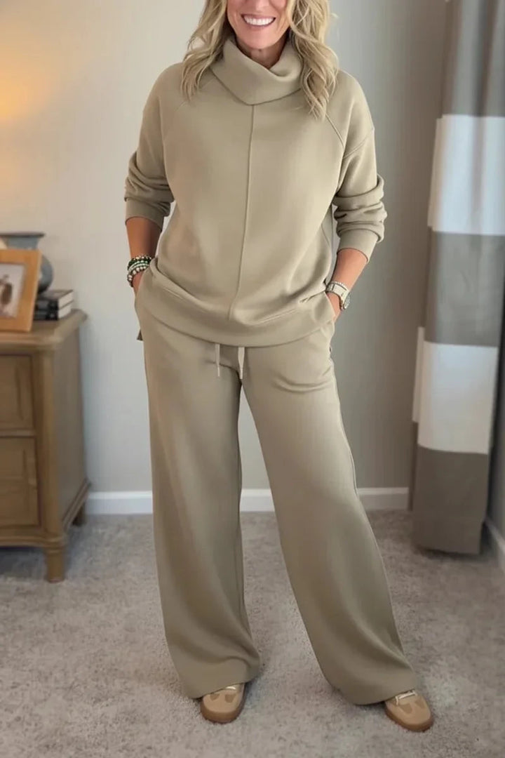 Woman wearing a khaki comfy set with long sleeves and wide-leg pants, standing in a carpeted room beside a wooden table.
