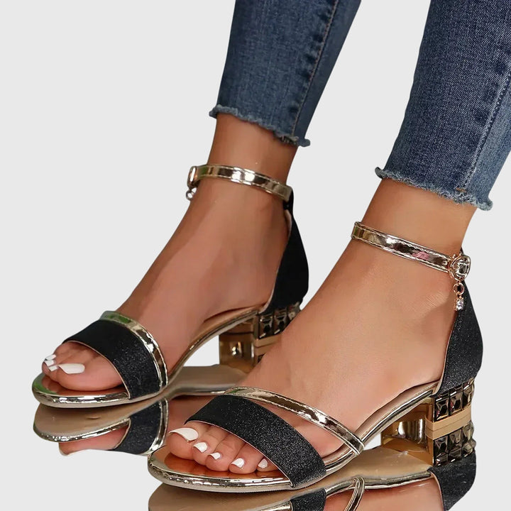 Black orthopedic heeled sandals featuring gold details and ankle strap, paired with frayed hem jeans, on a reflective surface.