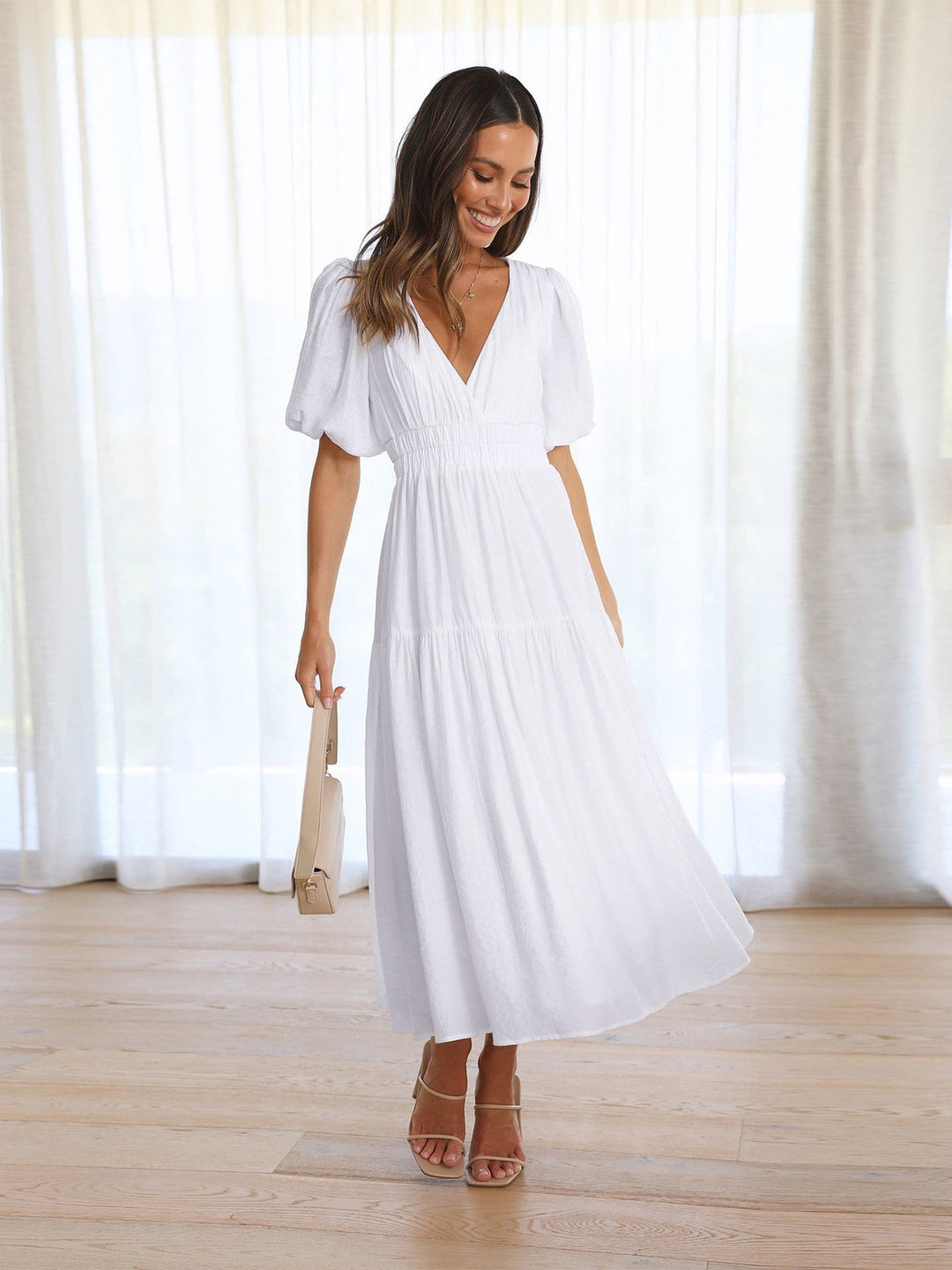 Woman in a white midi dress with puff sleeves, holding a beige bag in a bright room.
