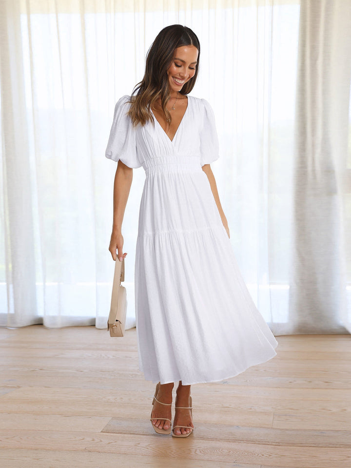Woman in a white midi dress with puff sleeves, holding a beige bag in a bright room.