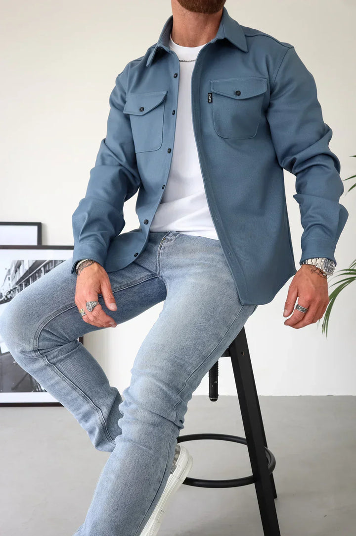 Blue buttoned overshirt with white t-shirt and light jeans, seated indoors.