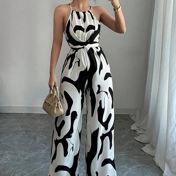 Model wearing a black and white printed backless jumpsuit, standing in a well-lit room with white curtains.
