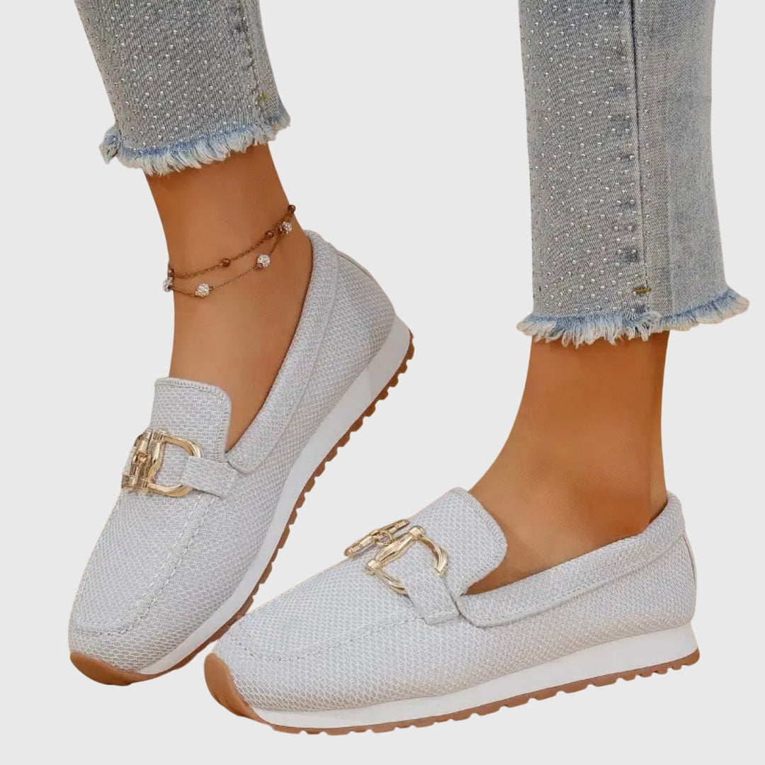 Light gray orthopedic loafers with gold accents worn with frayed jeans and anklet on a light background.