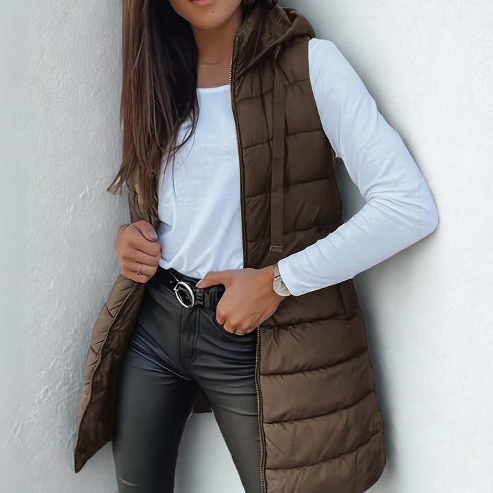Brown sleeveless puffer vest with hood over white top and black pants, white textured background.