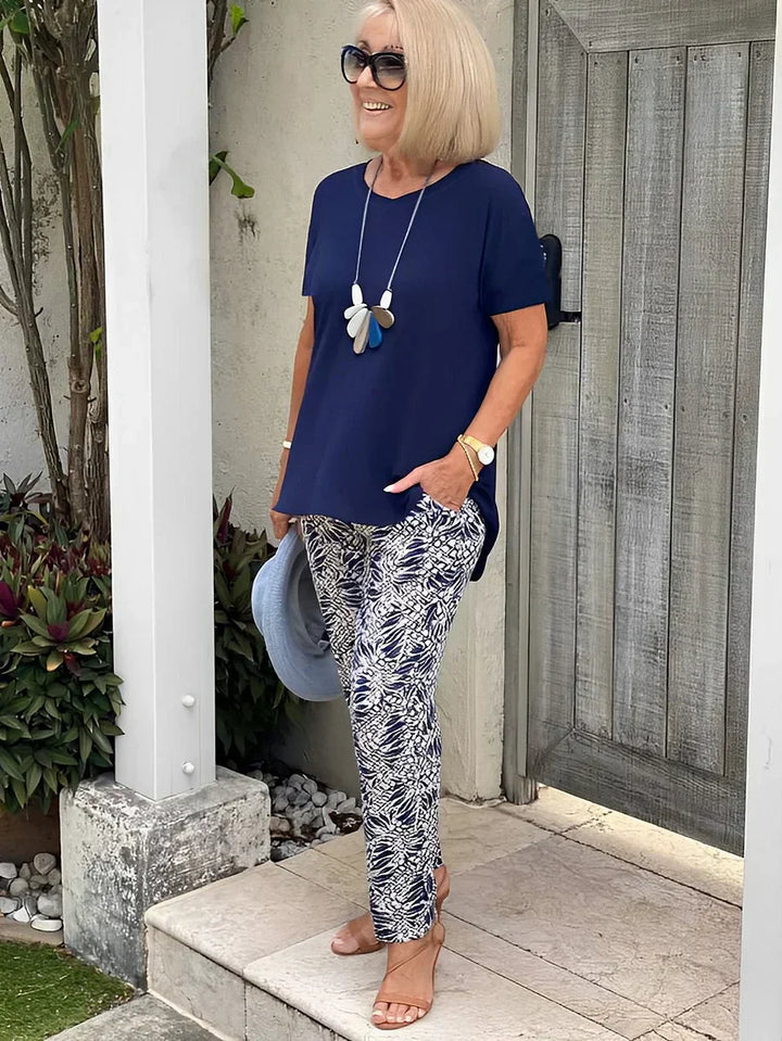 Woman wearing a navy loose two-piece set with patterned pants, accessorized with sunglasses and a necklace, holding a blue hat in a garden setting.