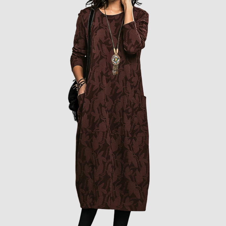 Woman in a stylish brown dress featuring a leafy design, complete with long sleeves and an ornate necklace, on a neutral backdrop.