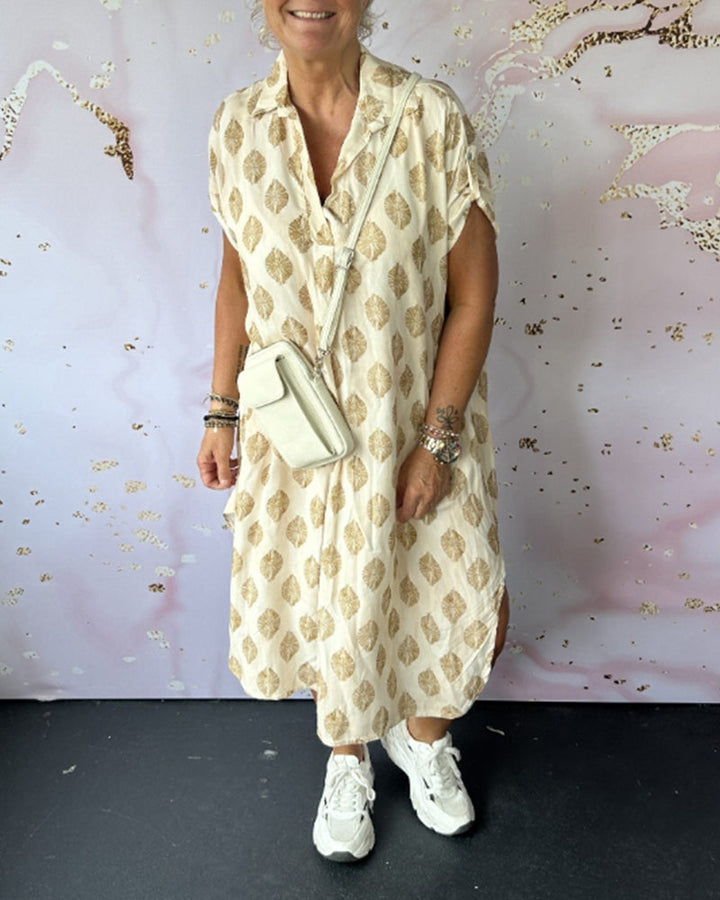 Model in beige dress with gold leaf designs, crossbody bag, white sneakers, against an abstract art background.