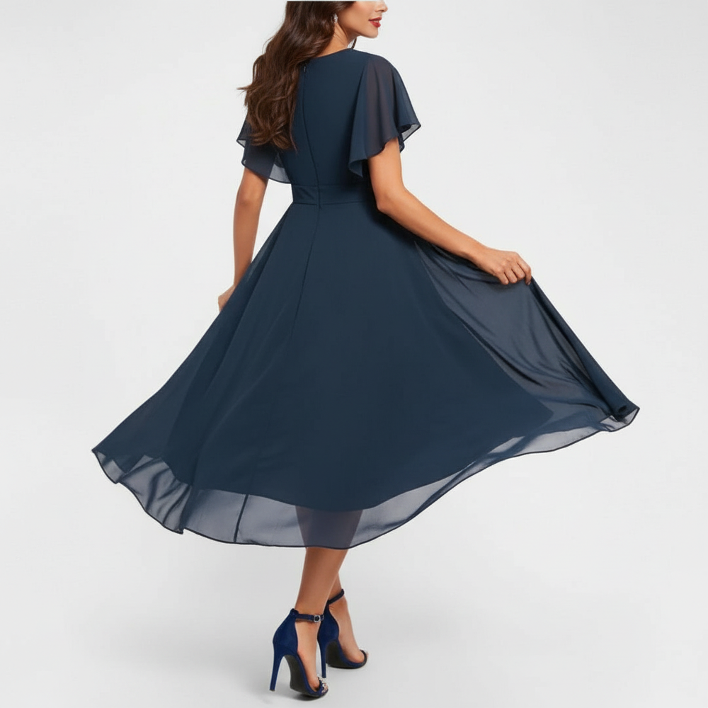 Back view of woman in navy flowing V-neck maxi dress, showcasing flared skirt and short sleeves, in a studio setting.