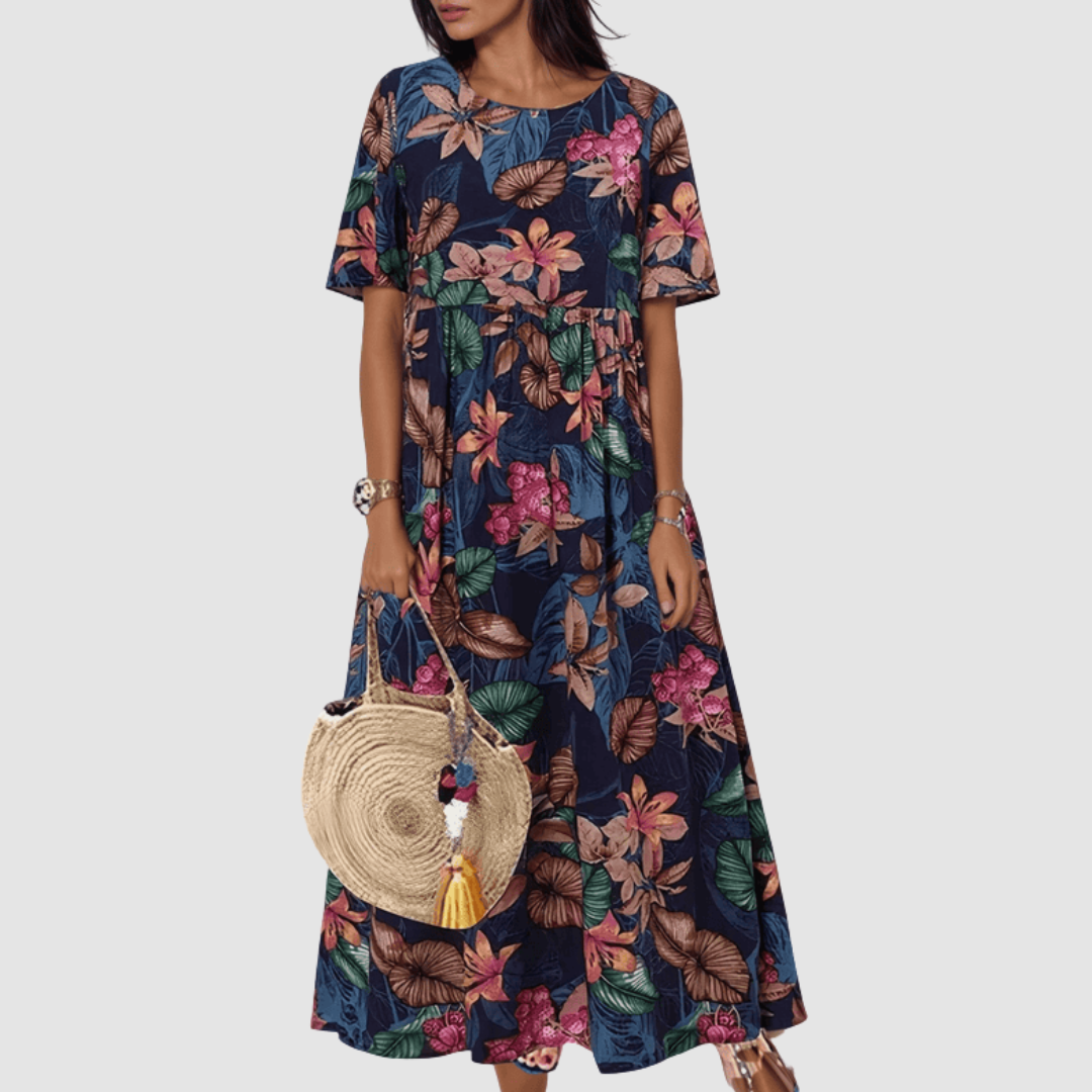 Model in a dark floral summer dress with vibrant patterns, holding a woven straw bag, on a plain background.
