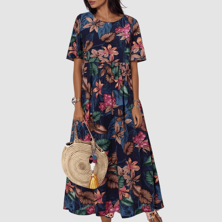 Model in a dark floral summer dress with vibrant patterns, holding a woven straw bag, on a plain background.