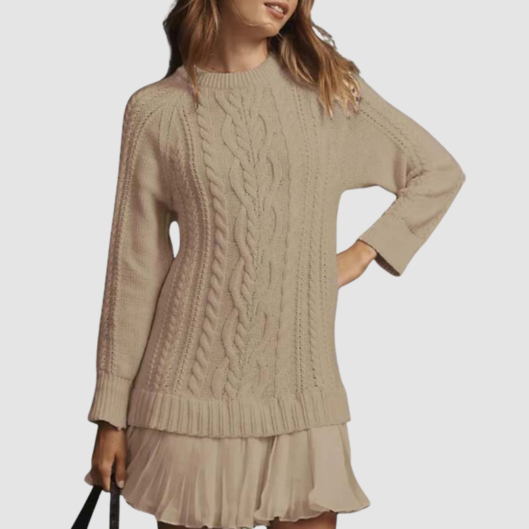 Beige cable knit mini dress with ribbed cuffs and a pleated hem, accessorized with a black handbag. Studio backdrop.