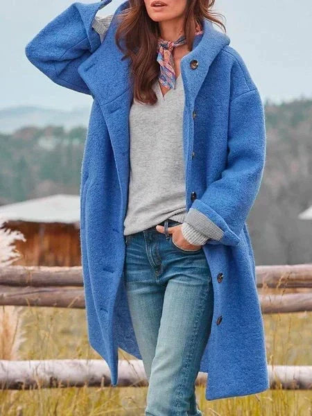 Woman in long blue coat with buttons, outdoors near a wooden fence with mountains in the background.
