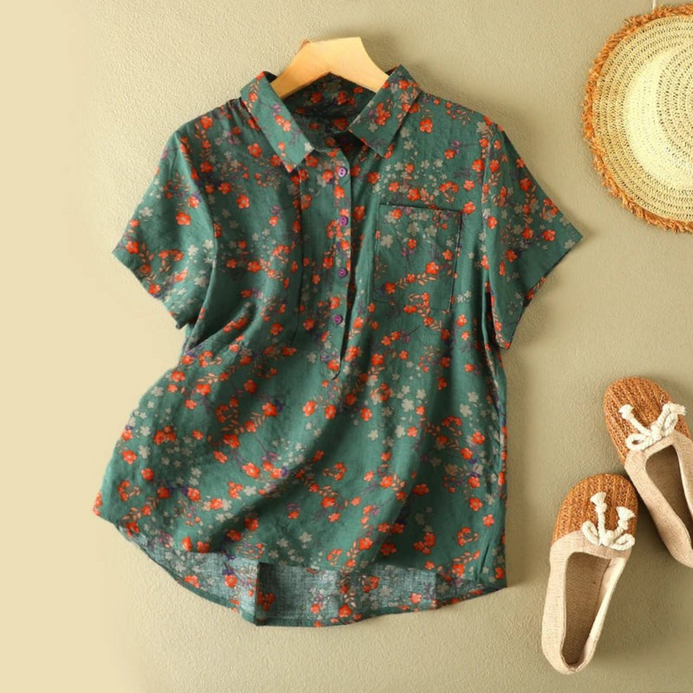 Green floral summer shirt with short sleeves, displayed on hanger against beige wall, next to straw hat and woven shoes.