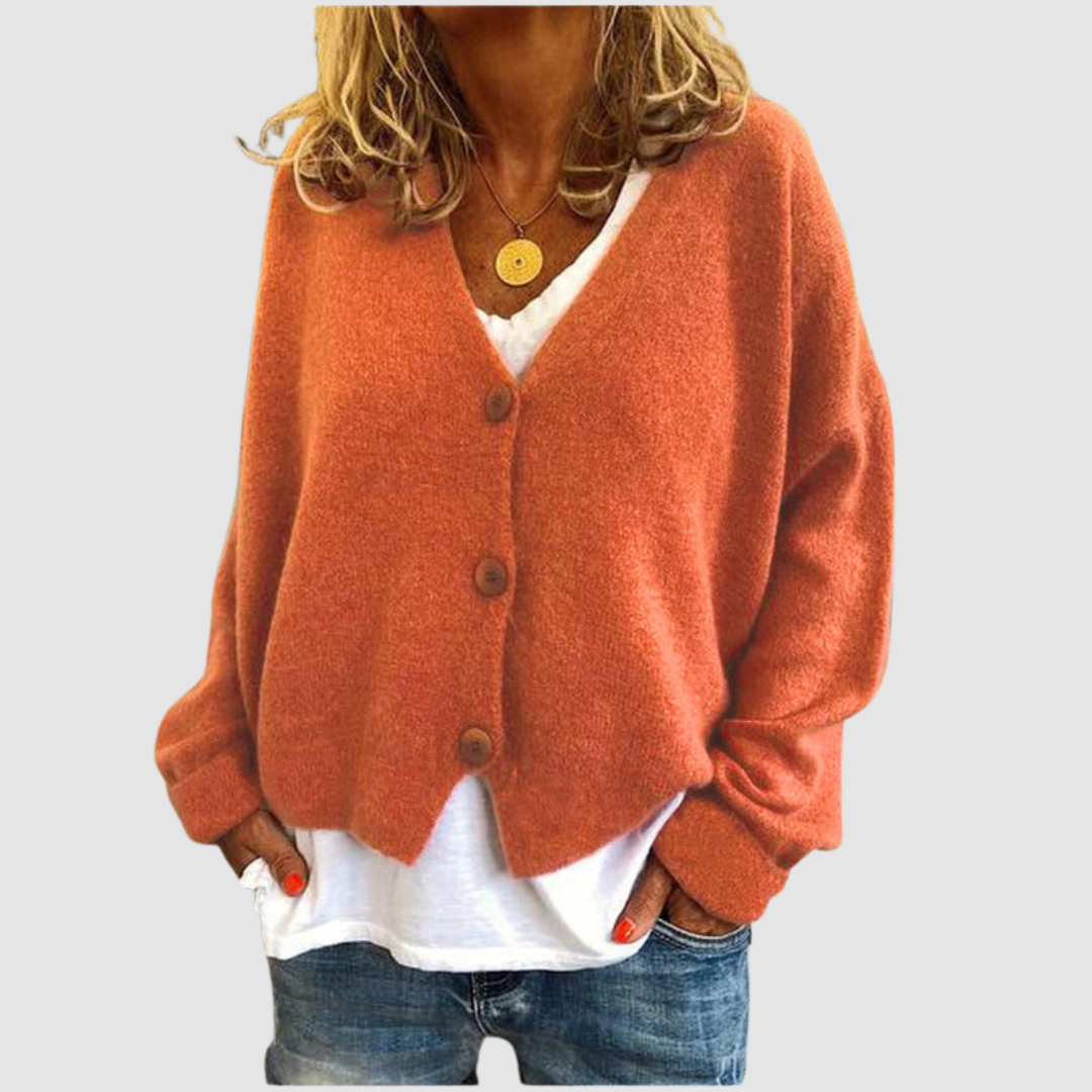 Woman wearing an orange knit cardigan over a white shirt and blue jeans, set against a plain gray background.