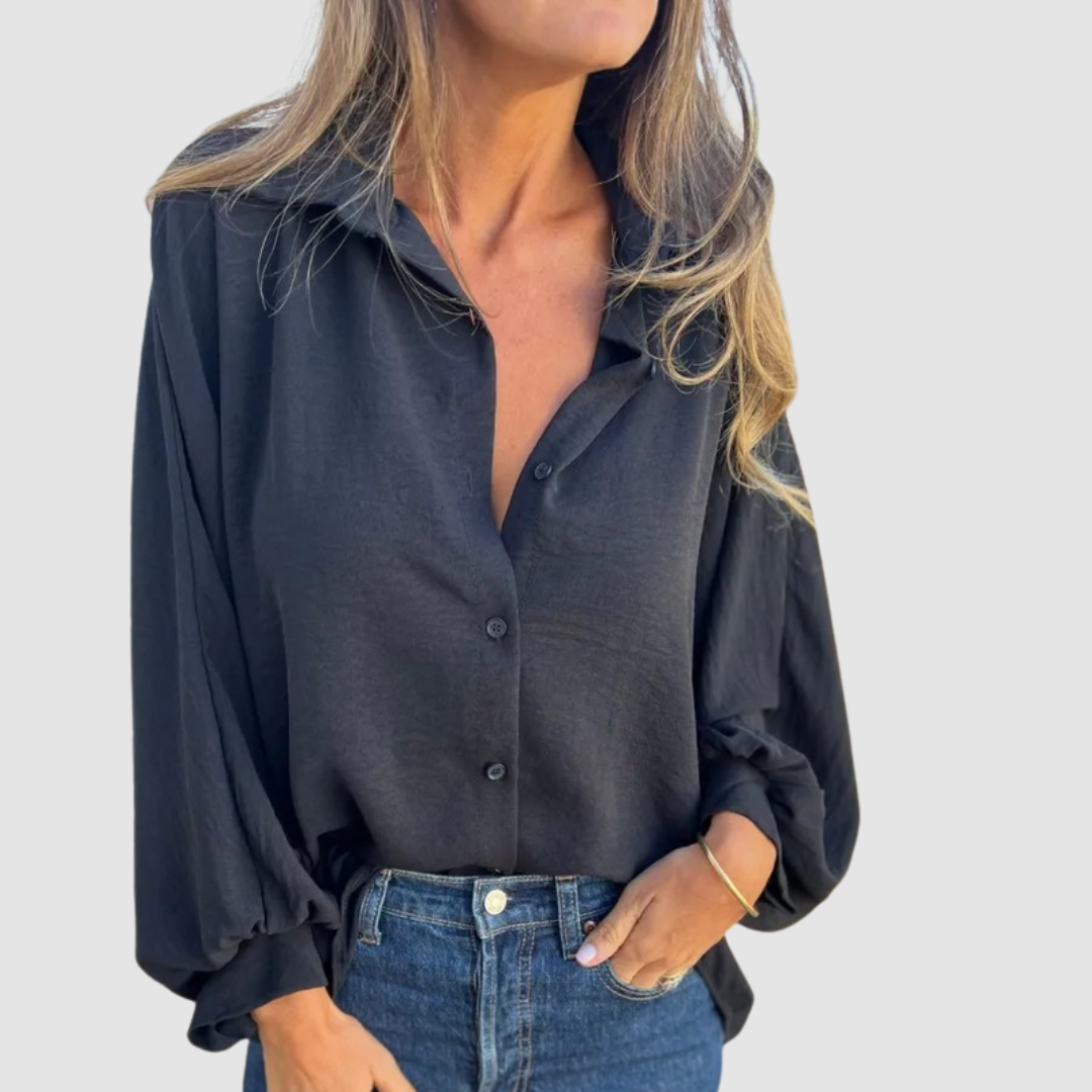 Model wearing a black oversized blouse with buttons, blue jeans, hand in pocket, gray background.