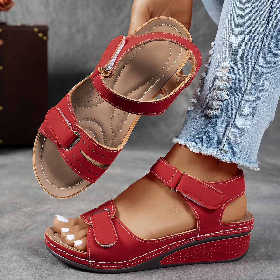 Red orthopedic sandal with dual straps and cushioned footbed, worn with ripped jeans on a gray stone floor.