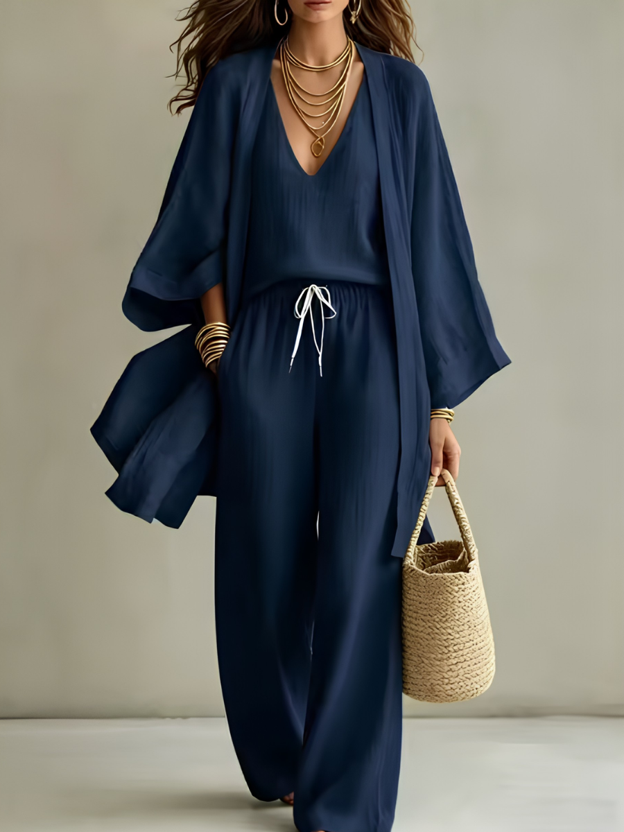 Navy three-piece relaxed homewear set with v-neck top, drawstring pants, flowy overcoat, and woven bag in studio setting.
