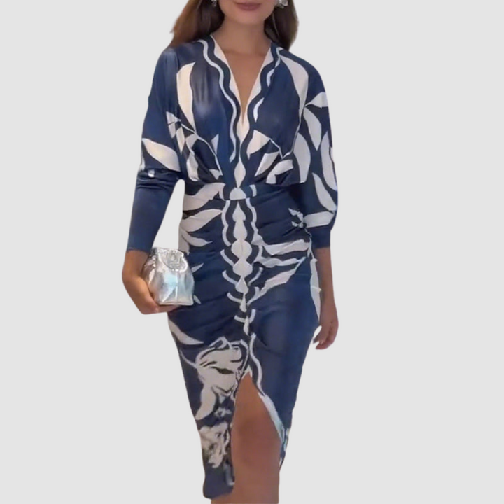 Stylish V-neck navy dress featuring white patterns, front slit, and long sleeves. Model accessorized with a silver clutch in a neutral studio.