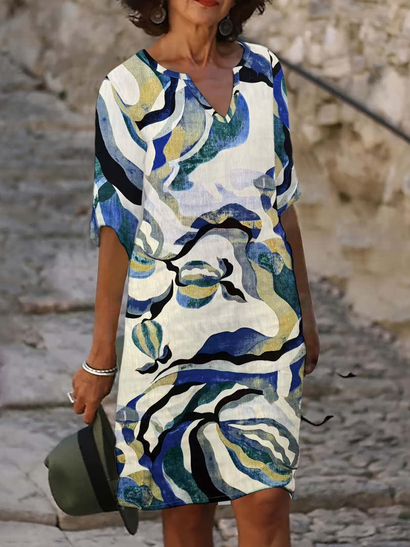 Woman in a colorful, abstract-patterned summer dress with blue, green, and yellow swirls, standing on stone stairs.