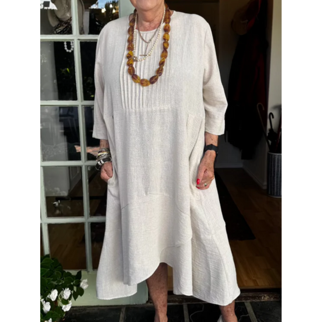 Woman wearing an off-white pleated dress with irregular hem, accessorized with necklaces in a boutique setting.