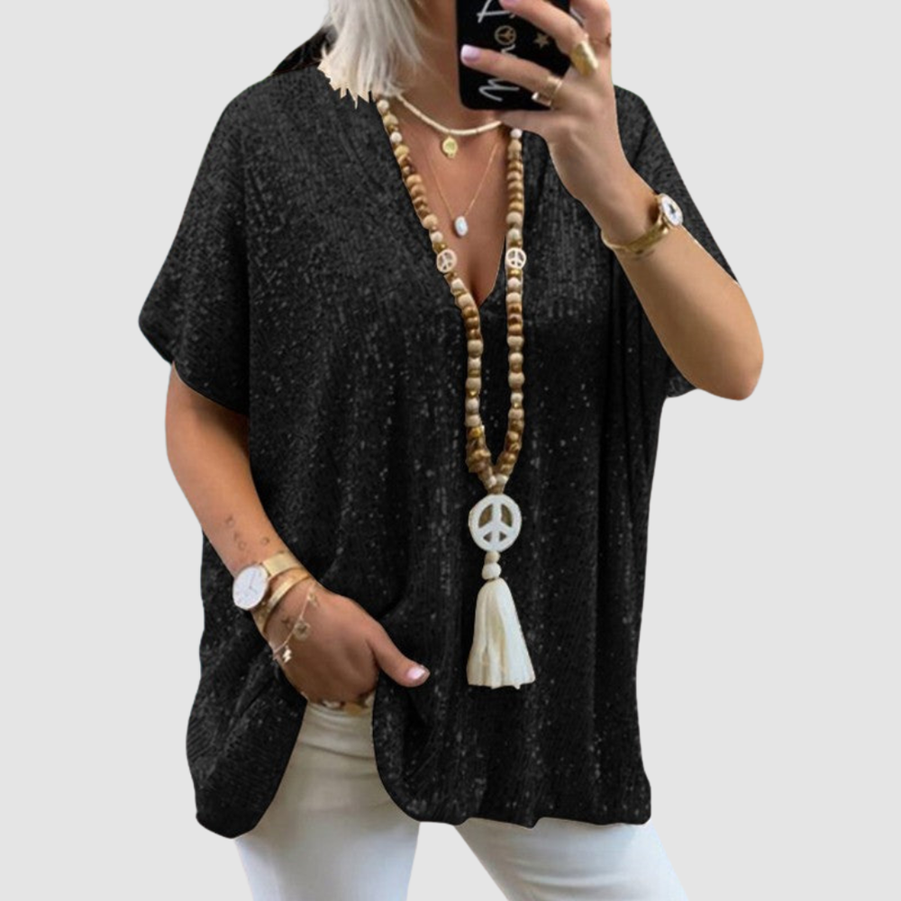 Woman wearing a black V-neck sequin top with white pants, accessorized with layered necklaces and bracelets, on a neutral background.