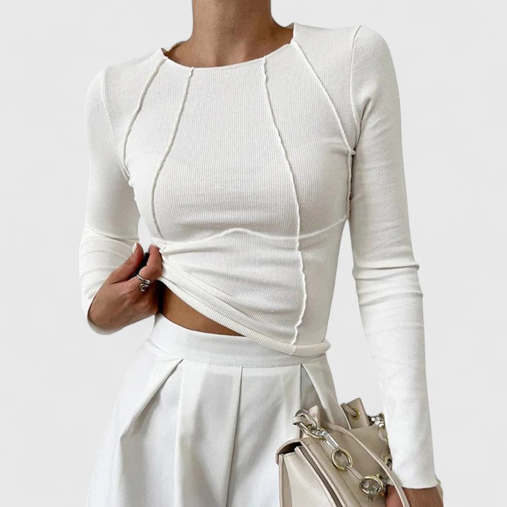 White long-sleeve top with curved seam details, worn with a white skirt and beige handbag, on a plain white background.