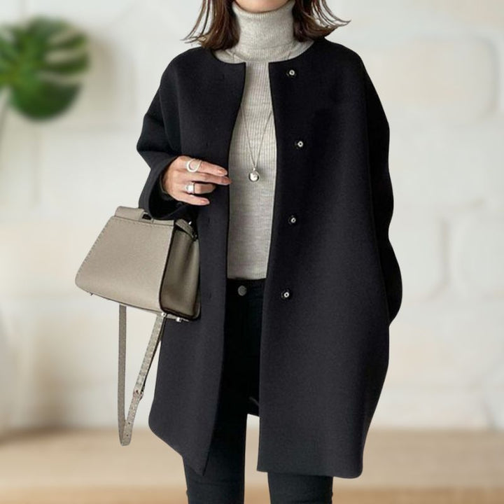 Black mid-length coat worn over a light turtleneck and black pants, styled with a beige handbag in a modern indoor setting.