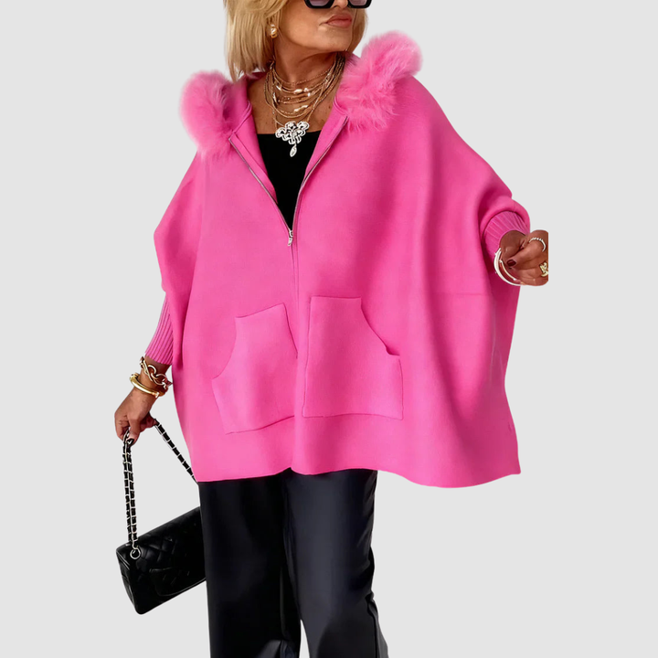 Woman wearing a bright pink casual coat with a fluffy hood, large pockets, and black pants, holding a black handbag in a studio setting.