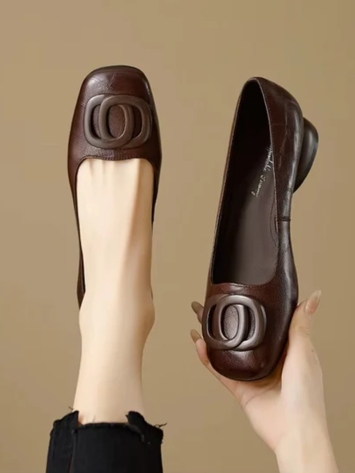 Brown leather flats with decorative buckle, one worn and one held, on a beige background.