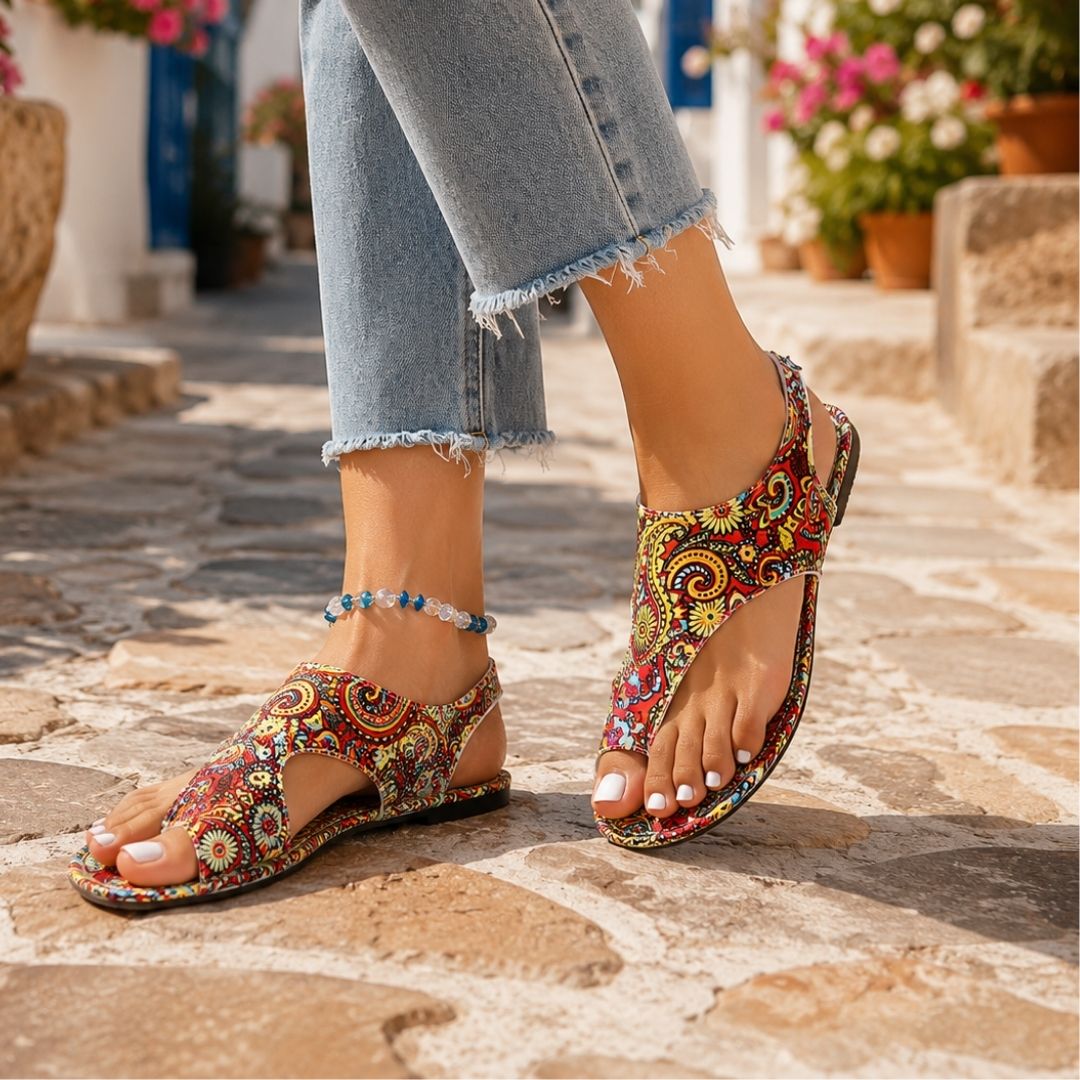 Colorful paisley-patterned orthopedic sandals on cobblestone path, person in denim jeans, outdoor setting with flowers.