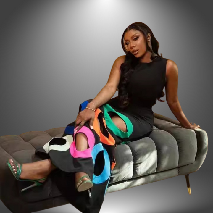 Person seated on a grey sofa wearing a black dress with colorful circular cut-outs in a studio environment.