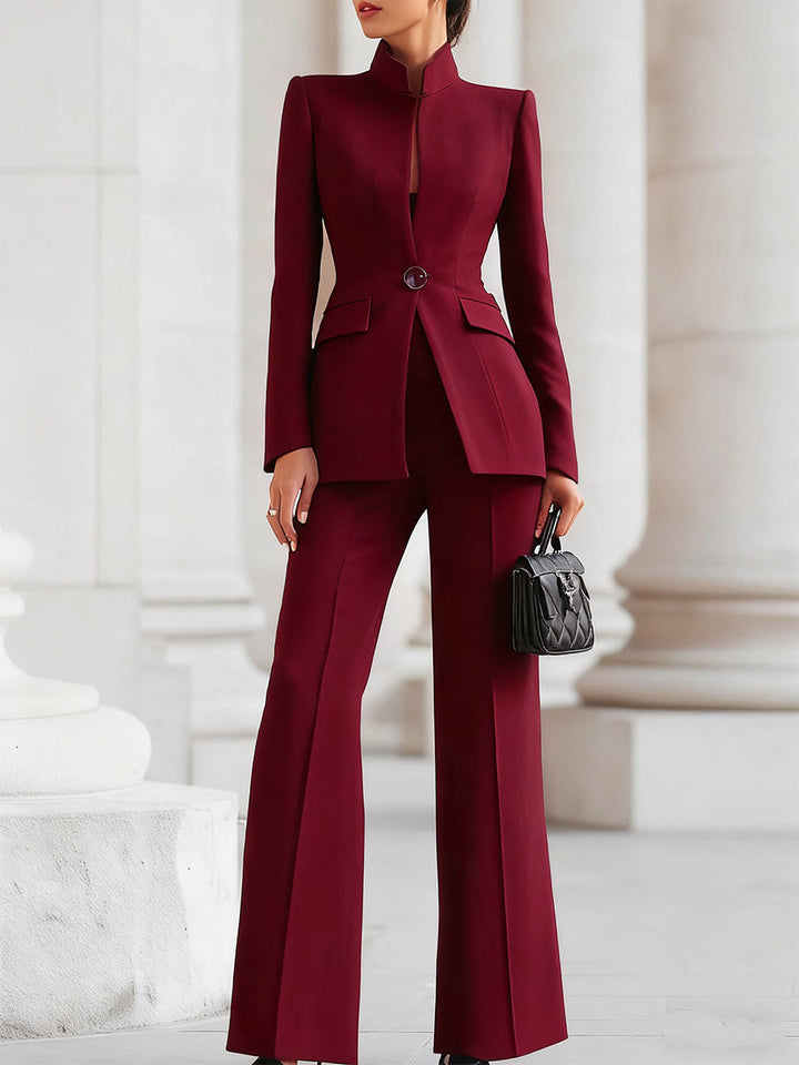 Elegant burgundy blazer and trouser suit set, worn outdoors with stone pillars in the background, person holding a black quilted handbag.