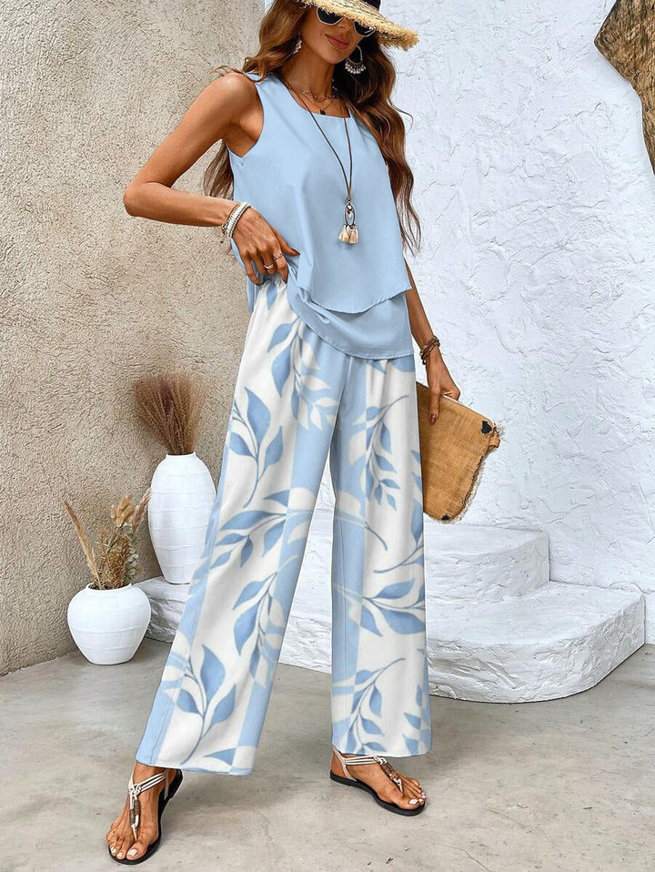 Woman in light blue top and leaf print pants, wearing a straw hat and sunglasses, standing by white steps with vases.
