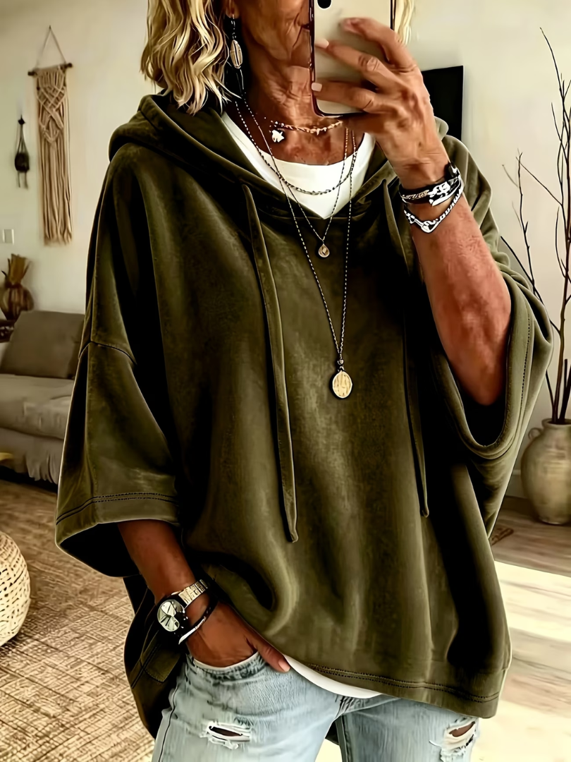 Olive green oversized velour hoodie with wide sleeves and drawstrings, worn over layered necklaces in a cozy, boho-styled living room.