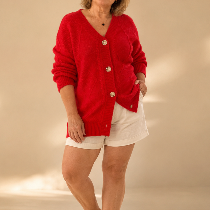 Person in a vibrant red knitted cardigan with buttons, paired with light shorts, standing in a softly lit beige environment.