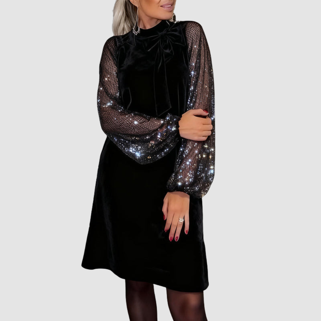 Black elegant dress with shimmering mesh sleeves, bow detail, and sequins, worn by a model, on a gray background.