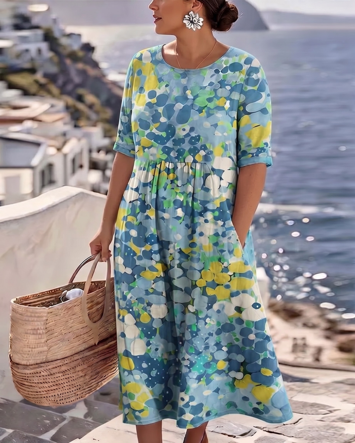 Focus on blue floral summer dress, model holding bag on terrace with ocean in background.