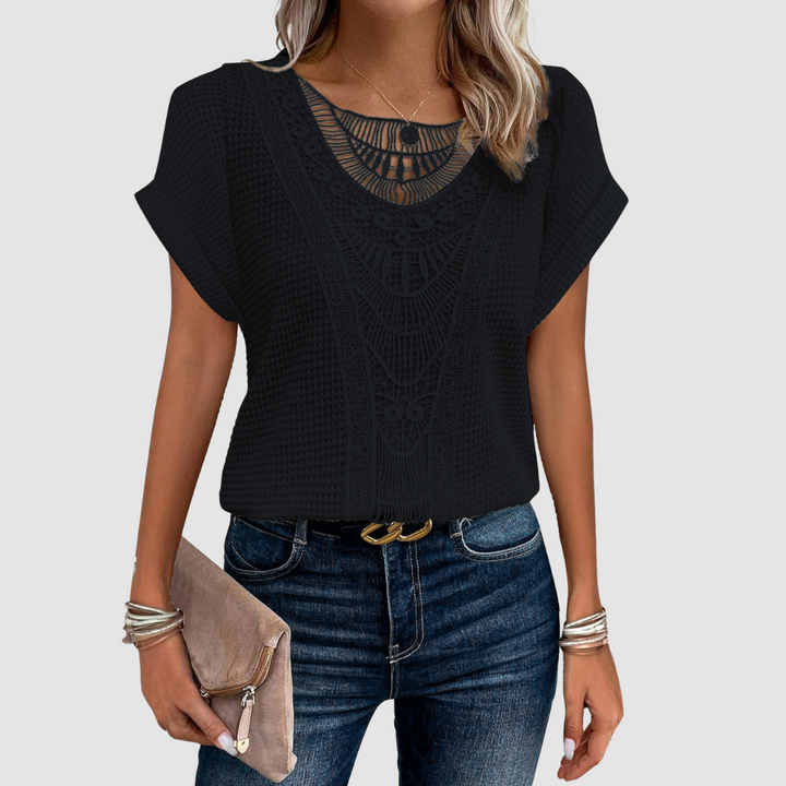 Woman wearing a black lace top with intricate patterns, paired with jeans, holding a beige clutch, studio background.