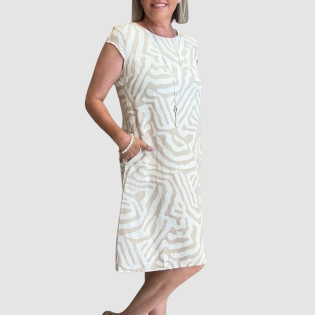 Woman in a light-toned, vintage-inspired dress featuring an abstract design, one hand in pocket, neutral background.