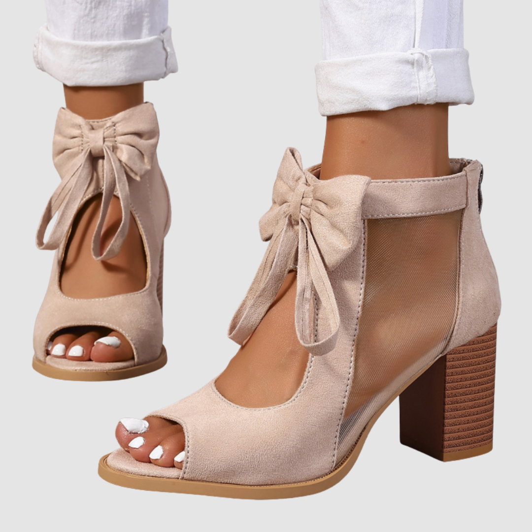 Side view of beige heeled sandals with bow and mesh design, paired with cuffed white pants on a neutral background.