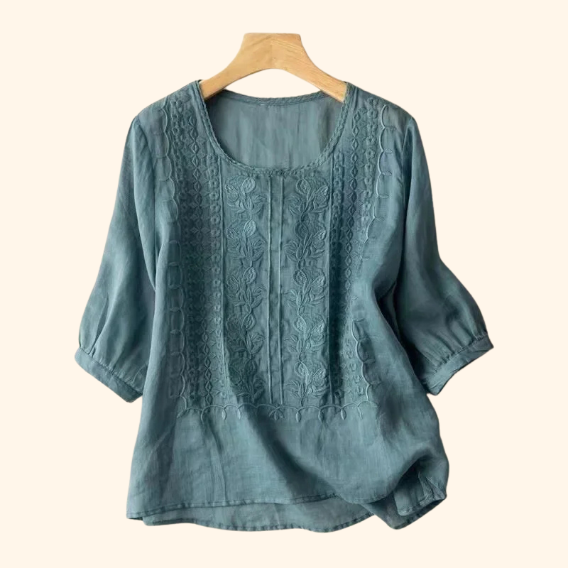 Teal elegant soft blouse with detailed embroidery, hung on a wooden hanger, light beige backdrop.