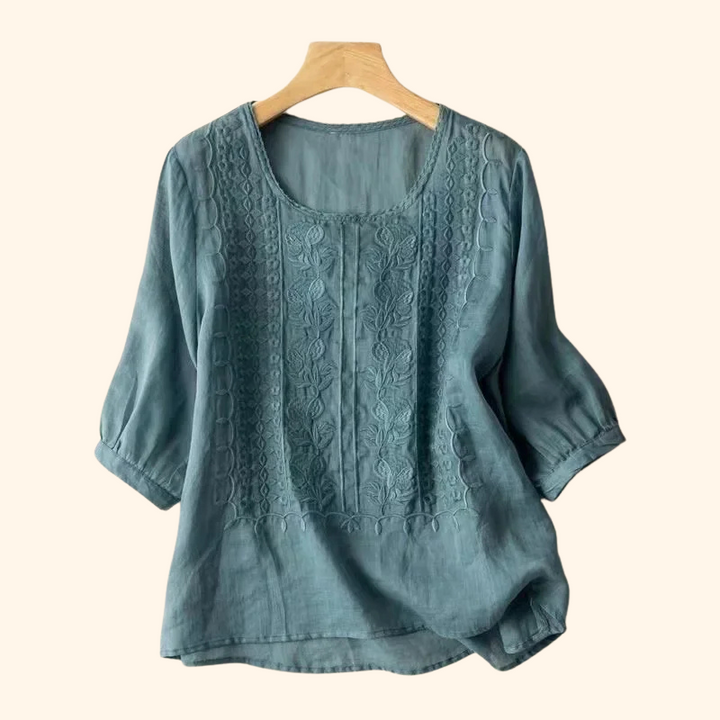 Teal elegant soft blouse with detailed embroidery, hung on a wooden hanger, light beige backdrop.